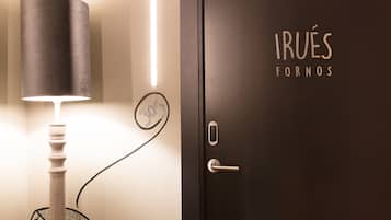 Una lámpara con pantalla cilíndrica, una puerta con el texto "IRUÉS FORNOS" y un logo circular, y una pared con un diseño geométrico.