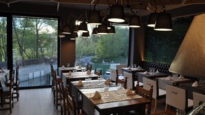 A restaurant with wooden tables, chairs, and hanging lamps. Large windows offer a view of trees and a road. - Alojamientos Ainsa Sanchez (Ainsa)