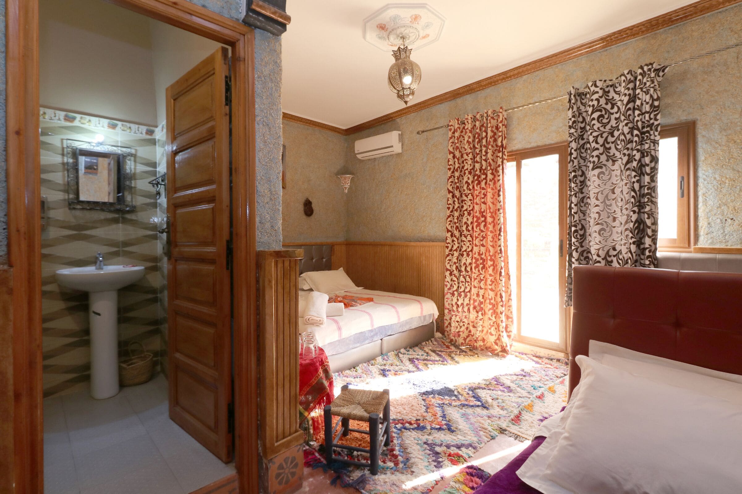 Un dormitorio con cama, mesita de noche, ventana con cortinas, aire acondicionado instalado en la pared y puerta que da a otra habitación.