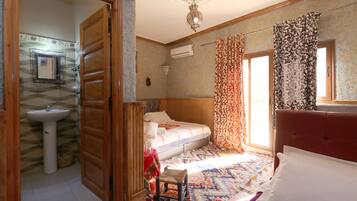 Un dormitorio con cama, mesita de noche, ventana con cortinas, aire acondicionado instalado en la pared y puerta que da a otra habitación.