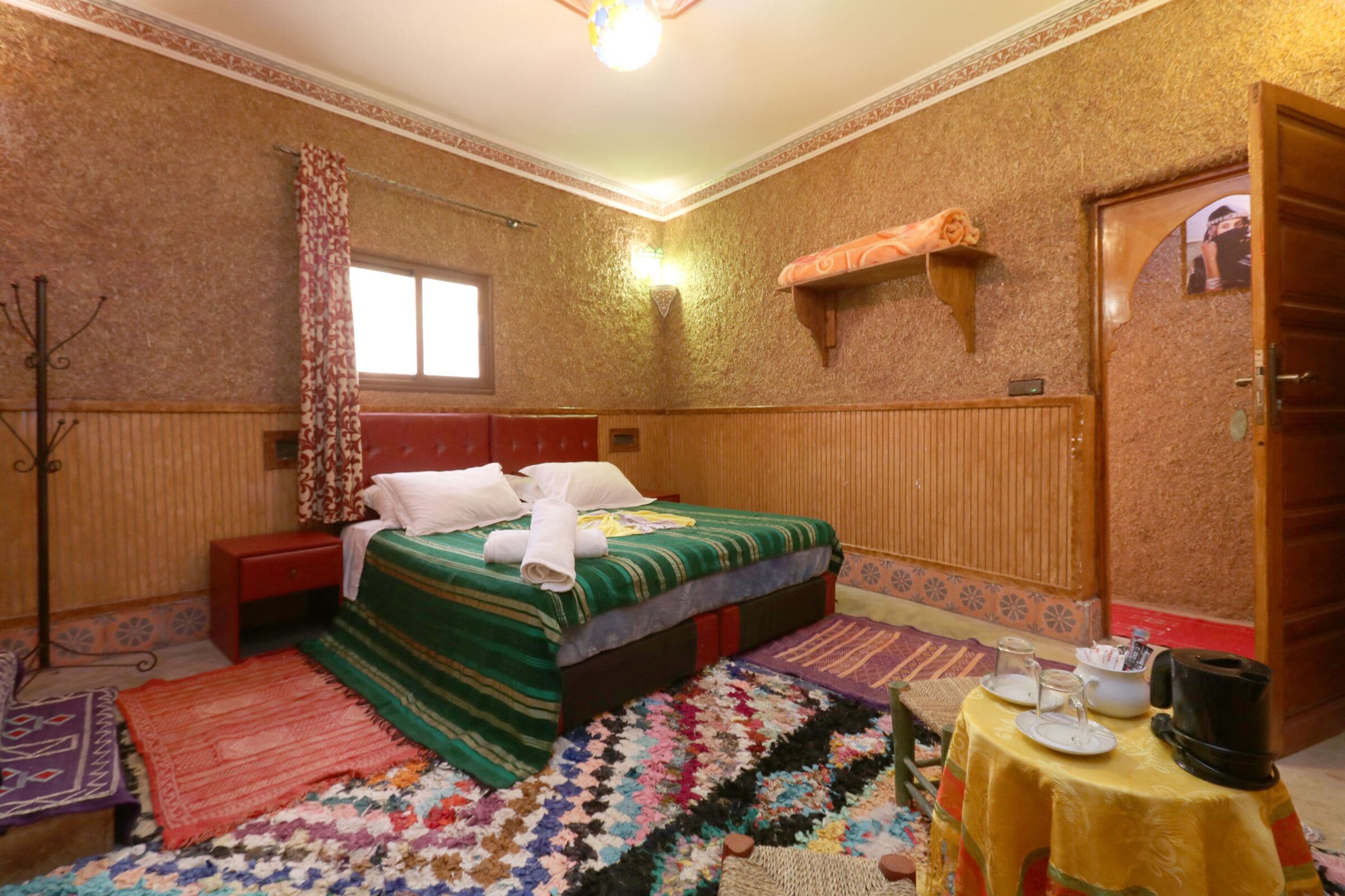 A hotel room with a bed, a small table with tea set, a red nightstand, a patterned rug, and a wooden shelf.
