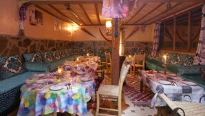 A cozy dining area with a stone wall, wooden ceiling, and a large sofa. Tables are set with plates, glasses, and candles. - Toubkal Garden Hotel (Asni)