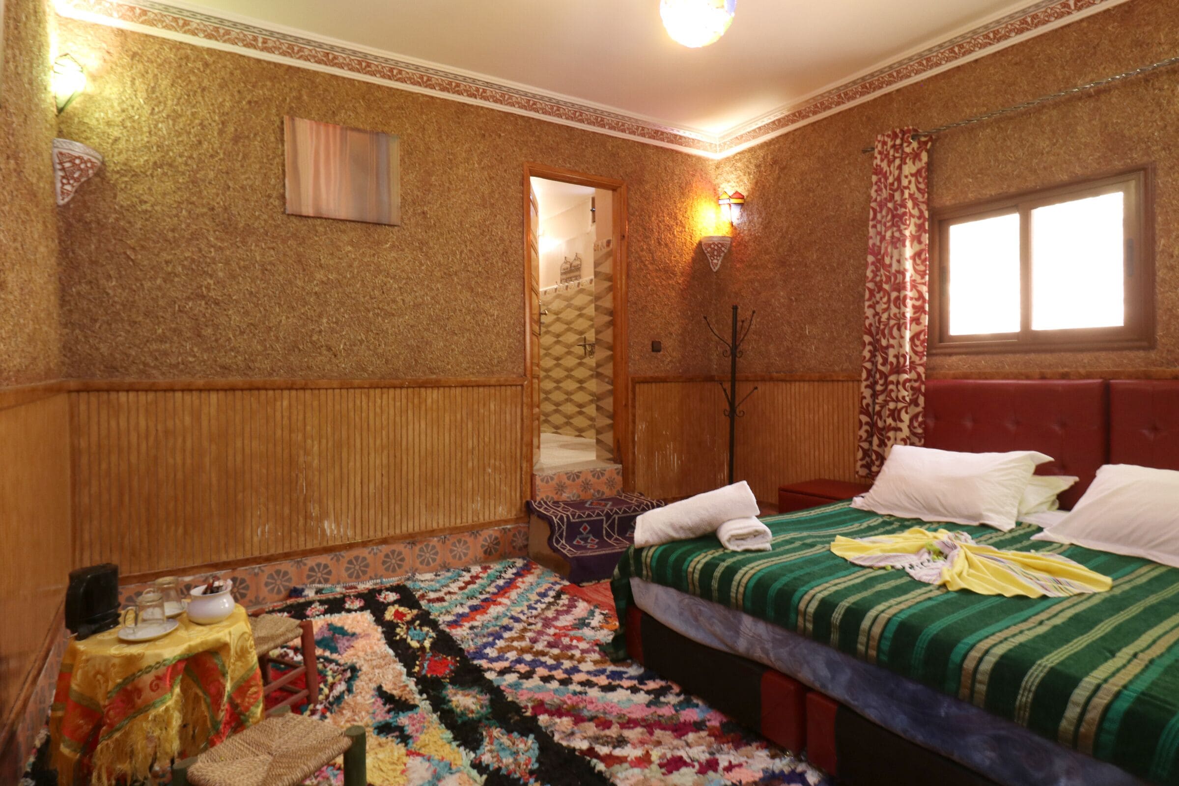 A hotel room with a bed, a small table with a tablecloth, a colorful rug, a wooden headboard, and a window with curtains.