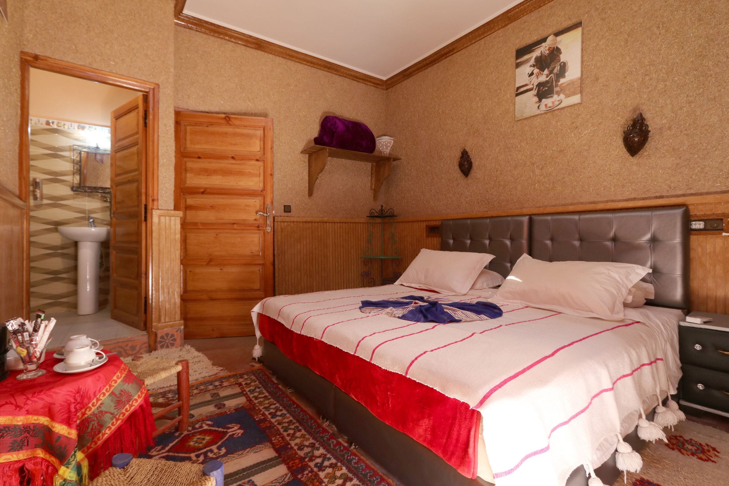 Un dormitorio con cama, cabecera de madera, mesita de noche, cómoda, espejo y estante con un objeto morado, además de una alfombra estampada.