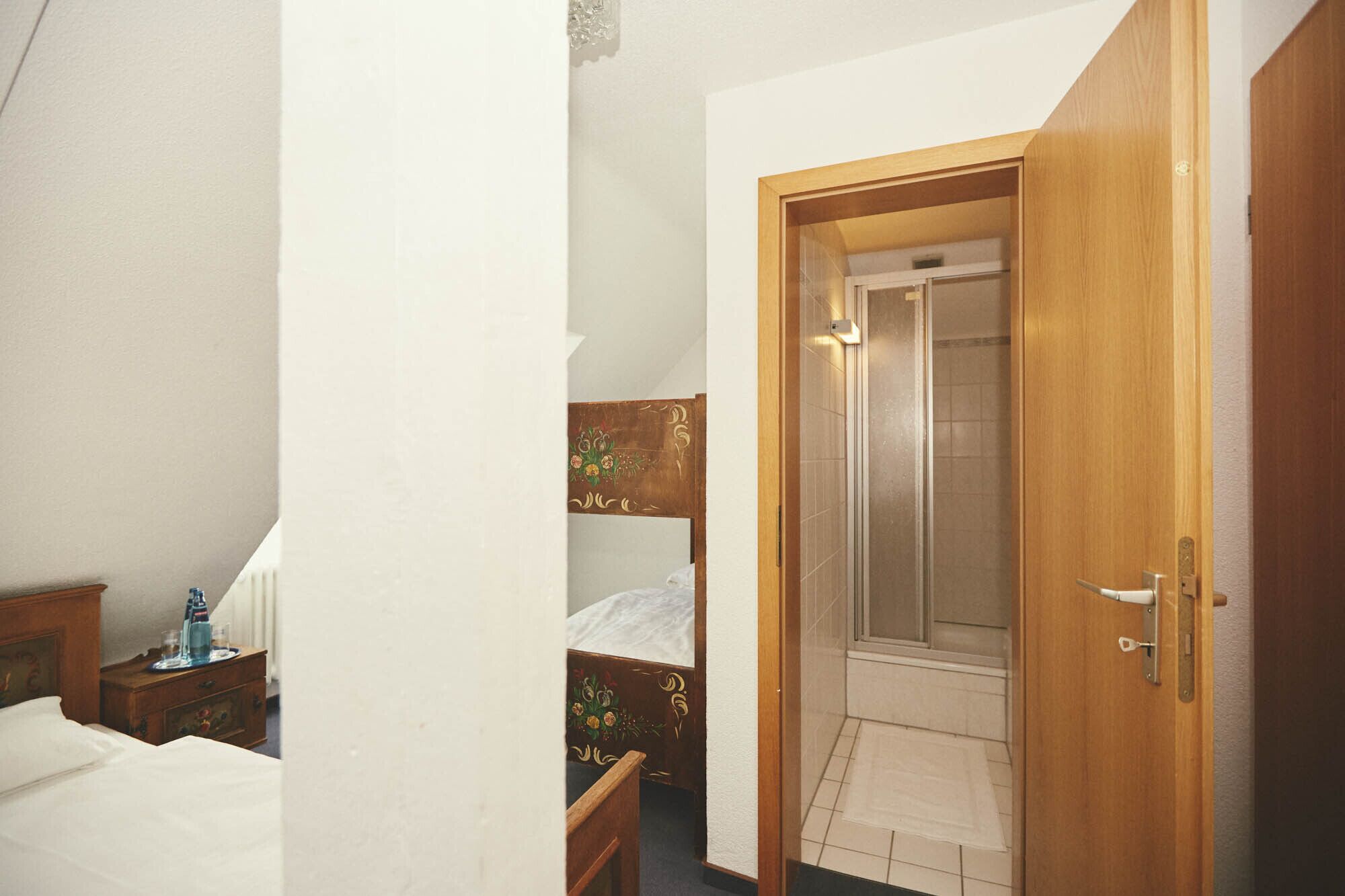 Habitació d'hotel amb un llit de fusta, una tauleta de nit, un bany amb porta de vidre i una petita cadira de fusta.
