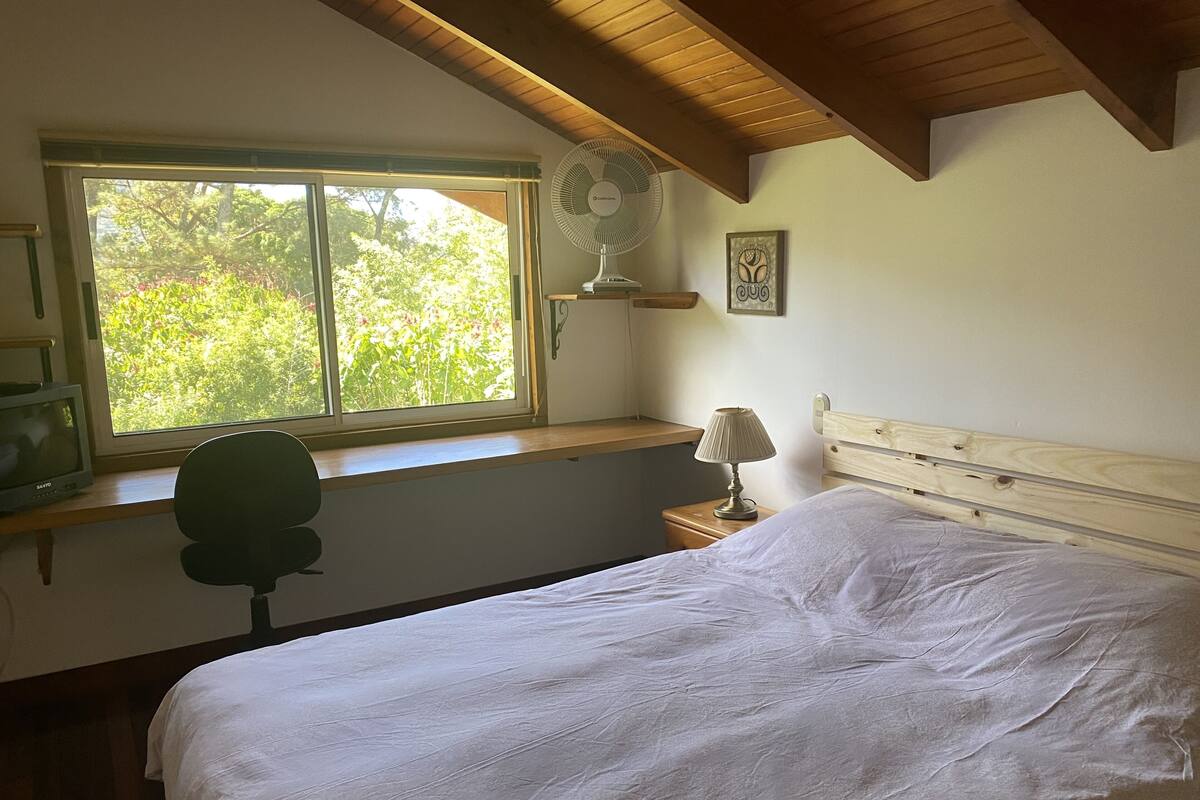 Une chambre avec un lit, un bureau avec une chaise, une fenêtre donnant sur un espace verdoyant, une lampe de chevet et un ventilateur fixé au mur.