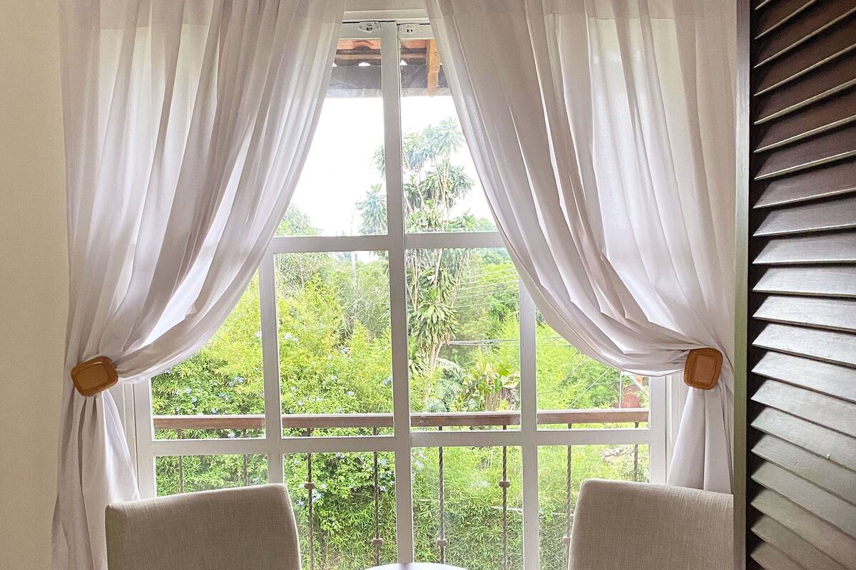 Une pièce avec deux chaises placées face à une petite table, une grande fenêtre avec des rideaux blancs et une vue sur la verdure à l’extérieur.