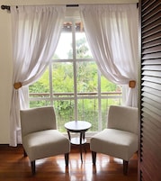 Habitación con dos sillas frente a una mesita, un ventanal con cortinas blancas y vistas a zonas verdes.