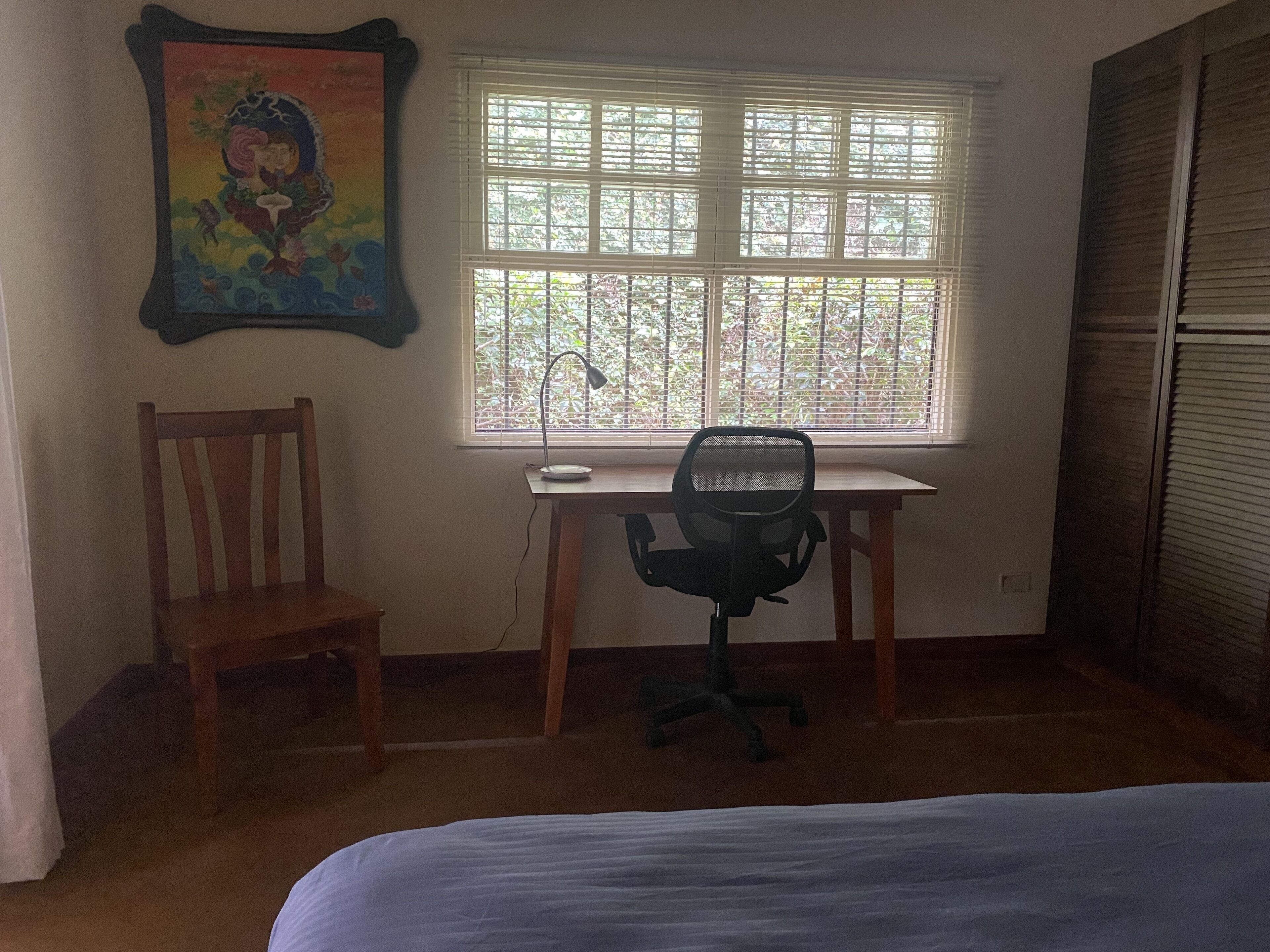 A bedroom with a bed, a desk with a chair, a window with blinds, and a painting on the wall.