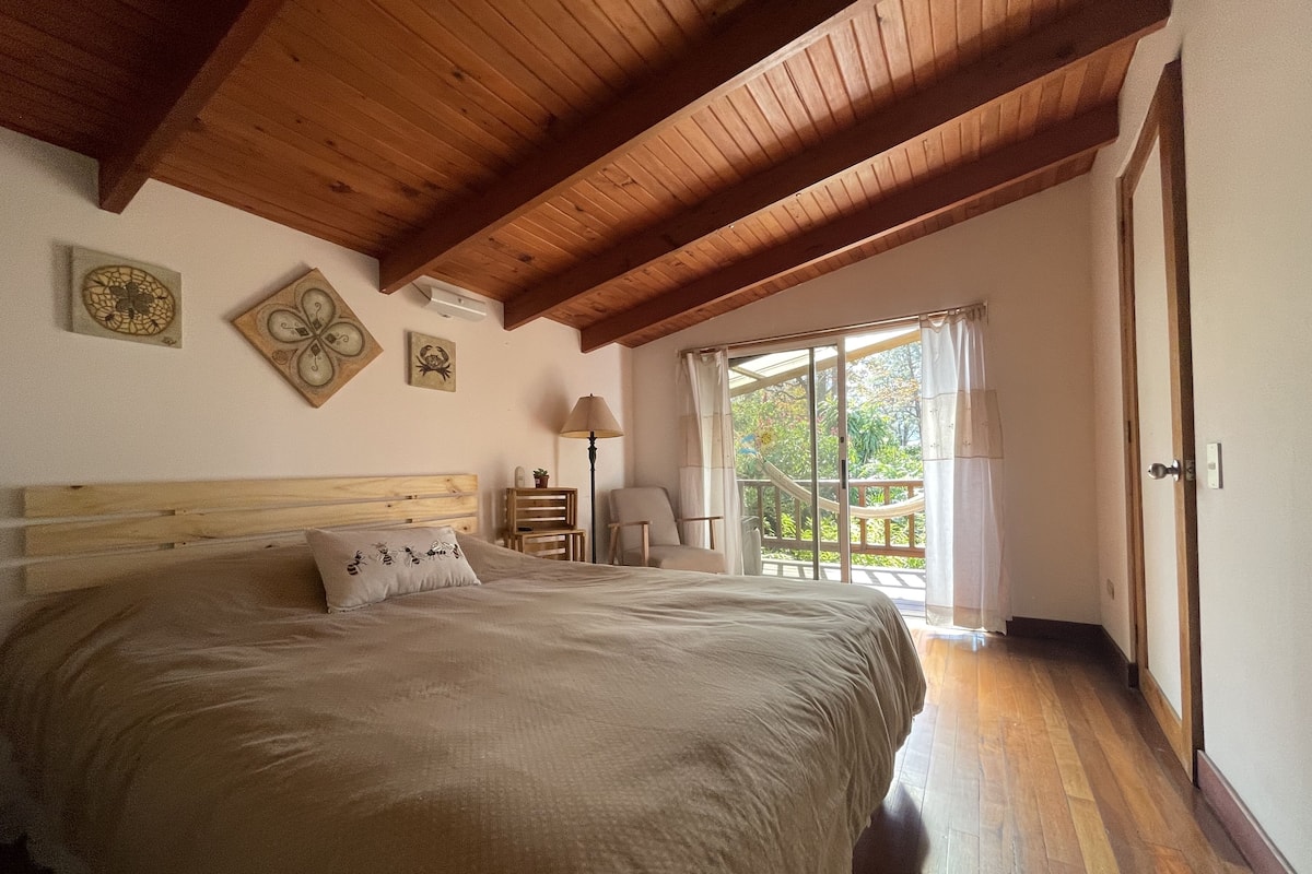 Une chambre avec un plafond en bois, un grand lit, une table de chevet, une chaise et une vue sur l’extérieur grâce à une porte coulissante.