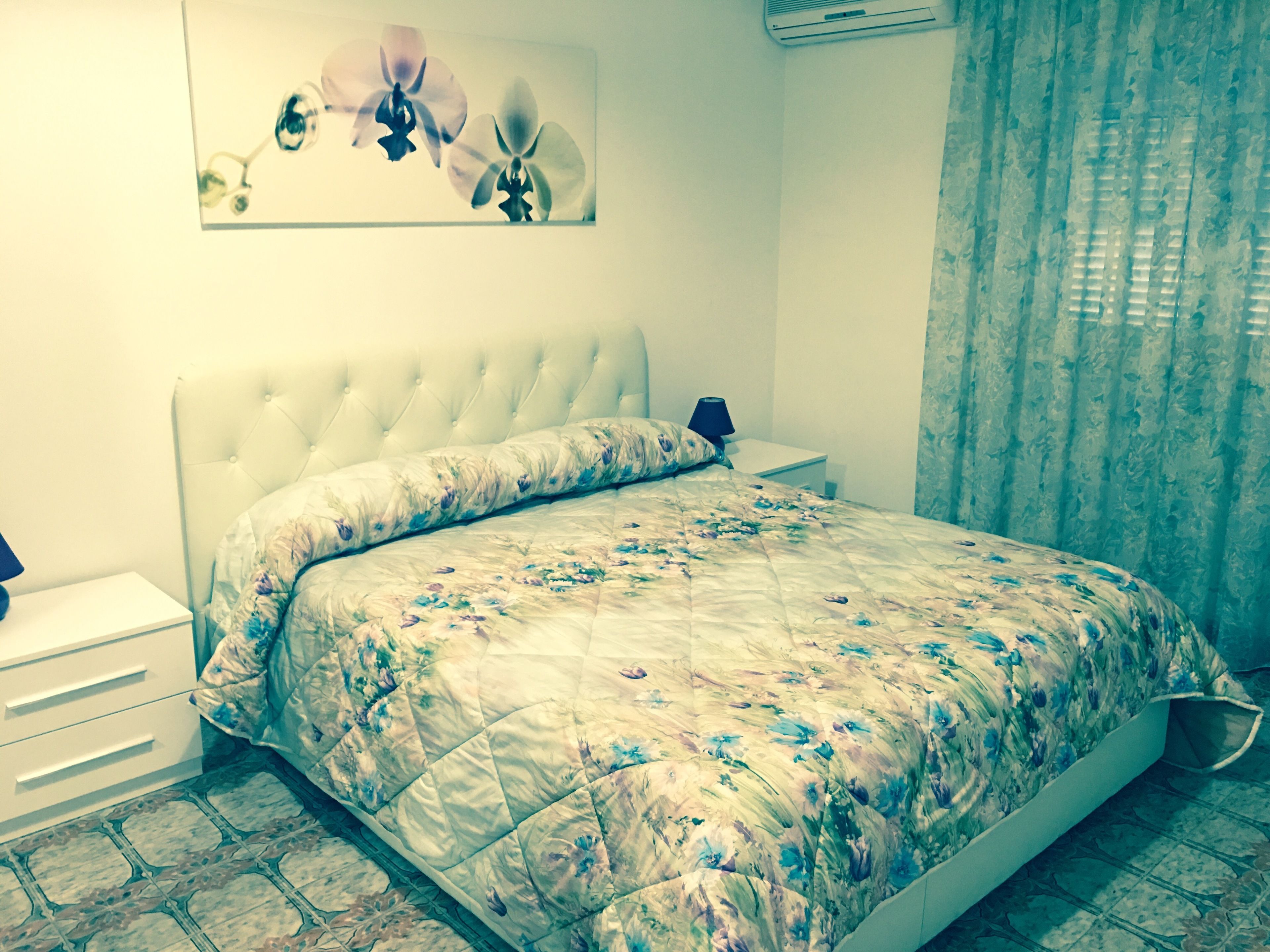 Un dormitorio con un cabecero capitonado, una colcha con estampado floral, una mesita de noche con lámpara y un aire acondicionado instalado en la pared.