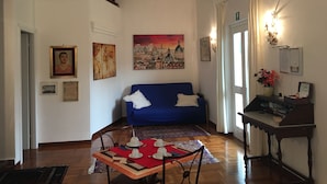 A living room with a blue sofa, a dining table with white cups and saucers, a wooden chair, a staircase, and a painting of a cityscape. - VillaGiò B&B (Frascati)