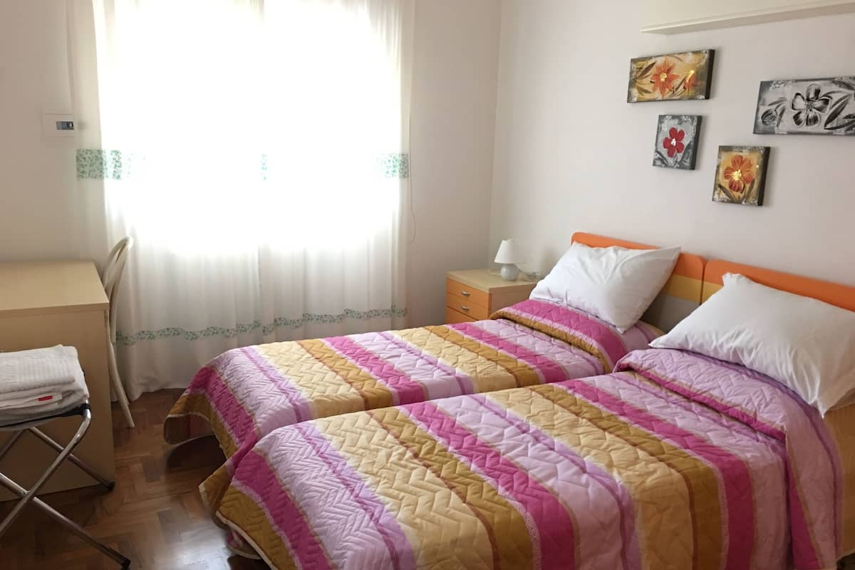 A bedroom with two beds, a desk, and a chair. There are wall-mounted paintings and a window with curtains.