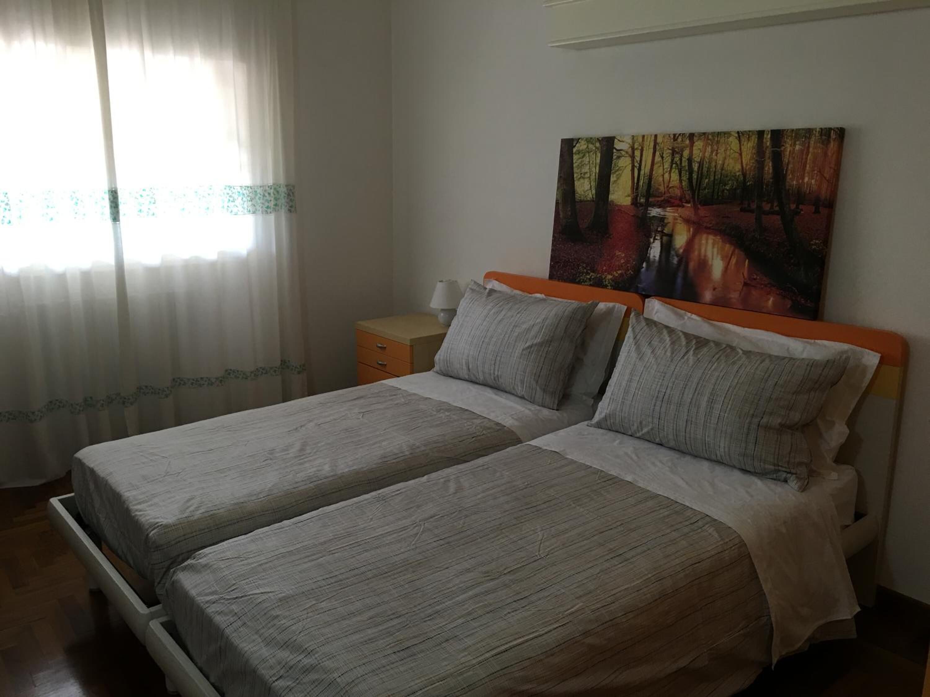 A double bed with a wooden headboard and two pillows, a nightstand with a lamp, and a painting of a forest scene on the wall.