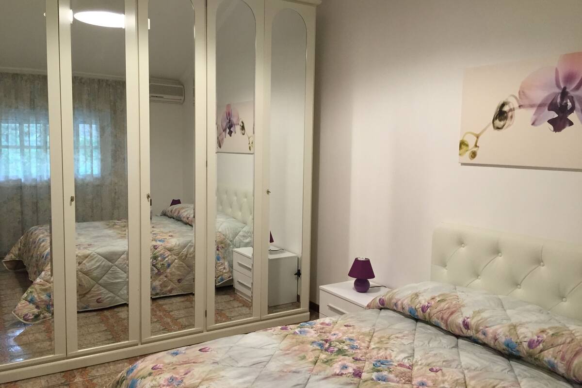 A bedroom with a large wardrobe, a bed with floral bedding, a bedside table, and a wall-mounted lamp.