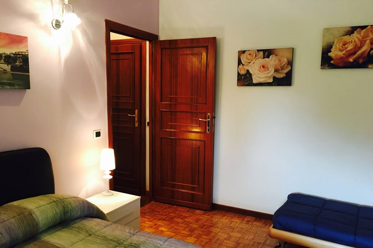 A bedroom with a bed, a bedside table with a lamp, a wooden door, and two paintings on the wall.