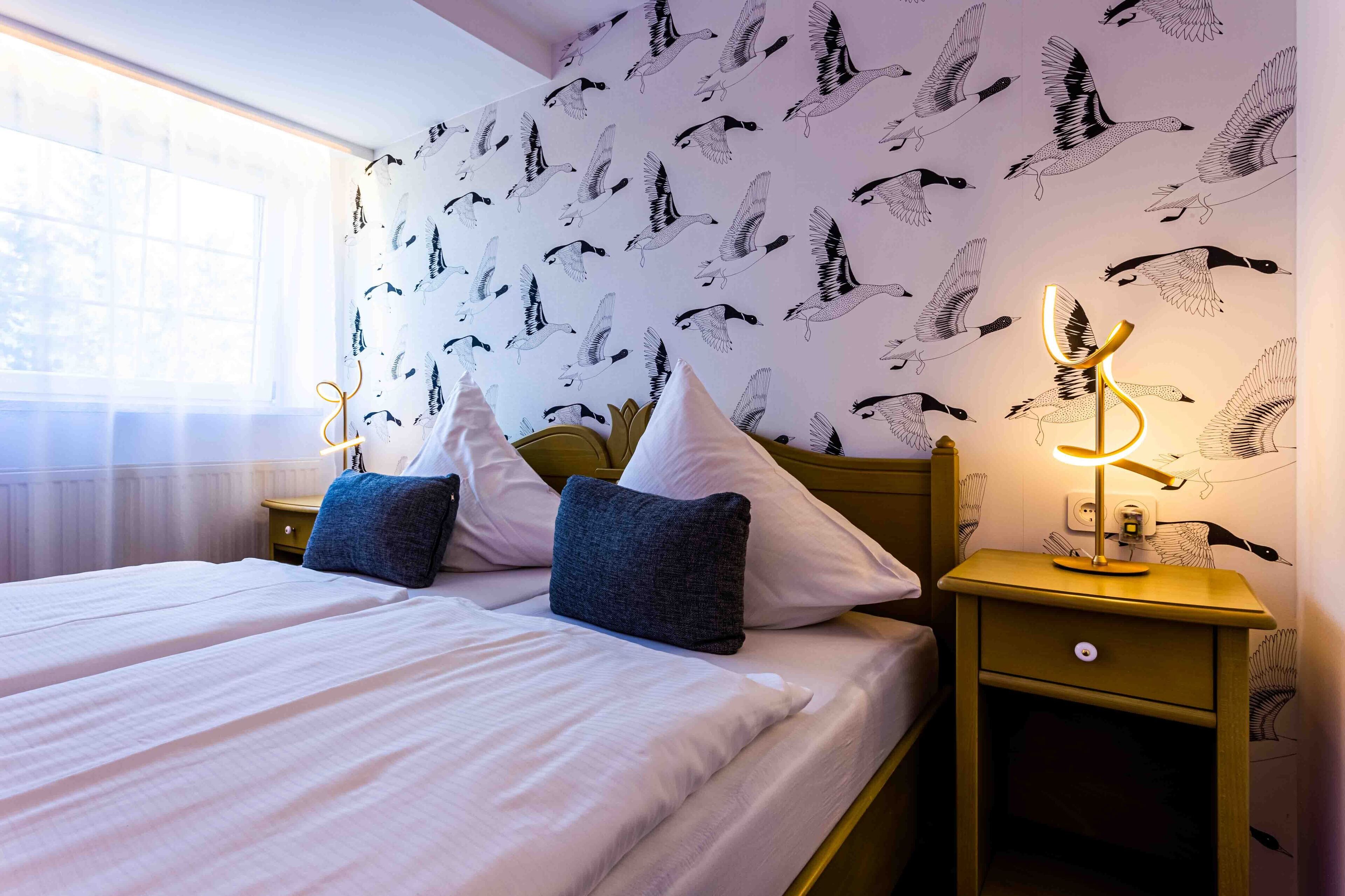 Ein Doppelbett mit weißen Leinen und zwei Kissen, ein Nachttisch mit Lampe sowie eine Wand mit Vogelmotiv.