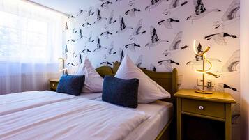 Ein Doppelbett mit weißen Leinen und zwei Kissen, ein Nachttisch mit Lampe sowie eine Wand mit Vogelmotiv.