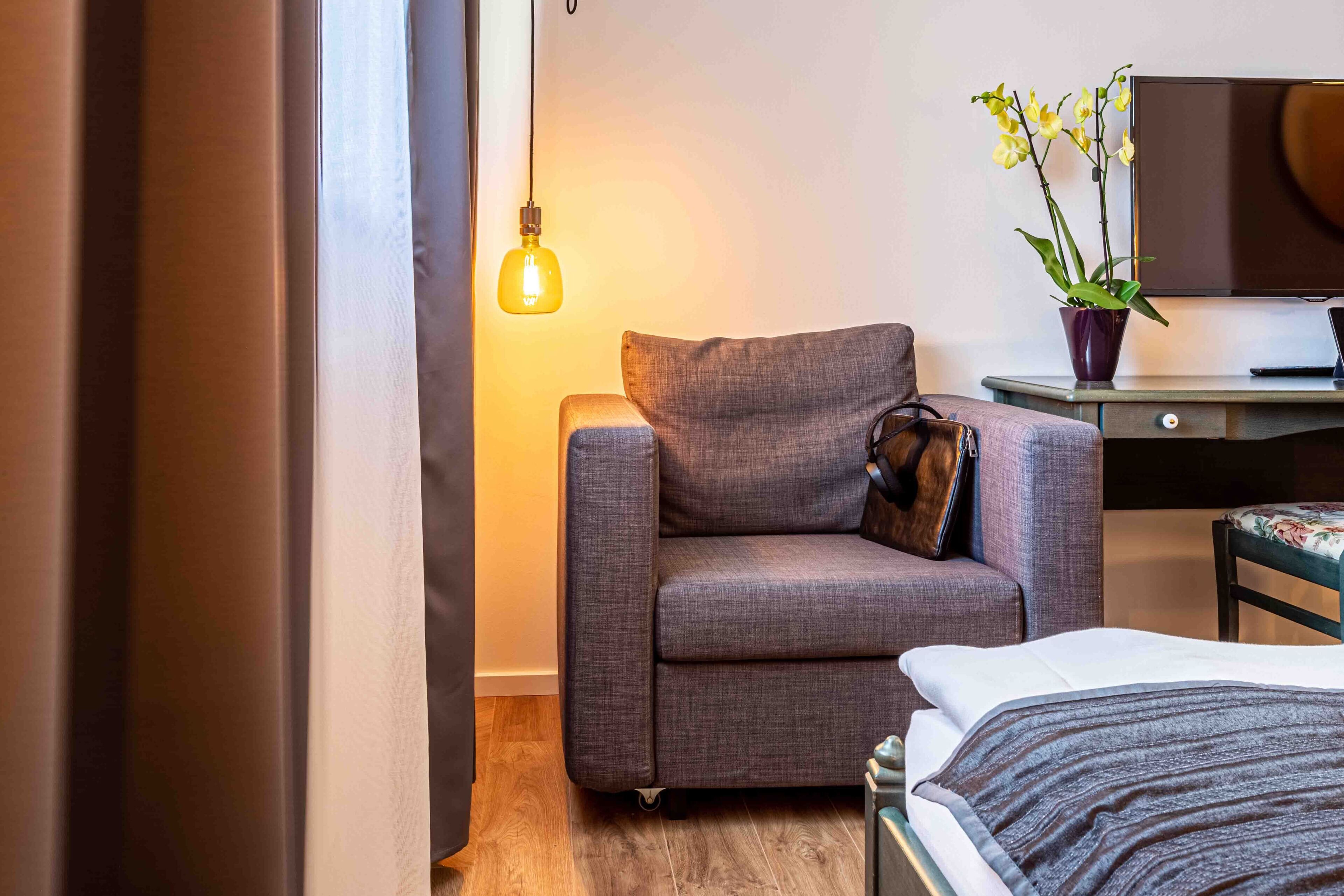 A modern hotel room with a grey sofa, a bed with a striped blanket, a desk with a laptop, a potted orchid, and a hanging light fixture.
