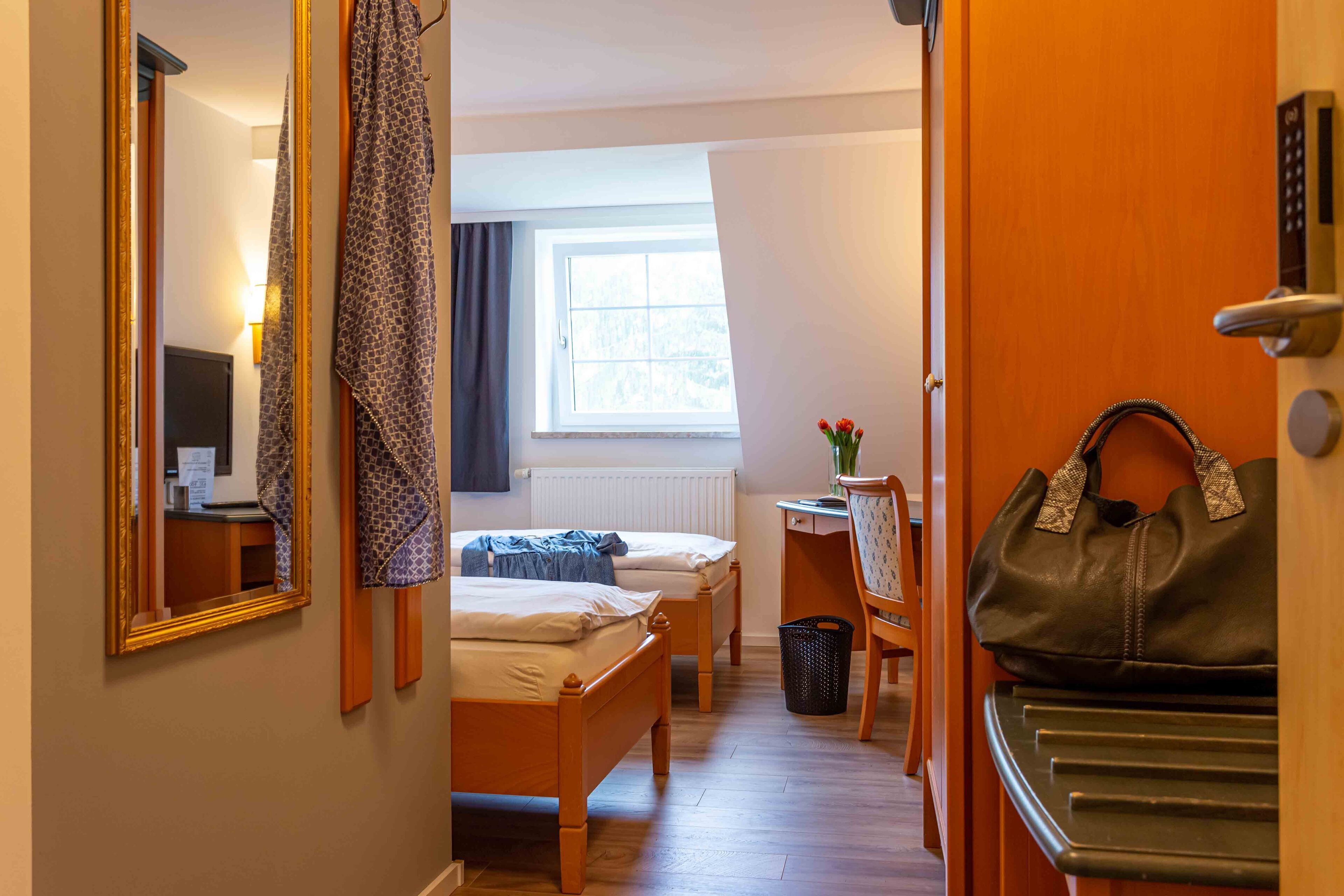 A hotel room with a wooden bed, a desk with a chair, a window with curtains, a mirror, and a handbag on a stand.