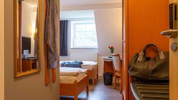 A hotel room with a wooden bed, a desk with a chair, a window with curtains, a mirror, and a handbag on a stand.