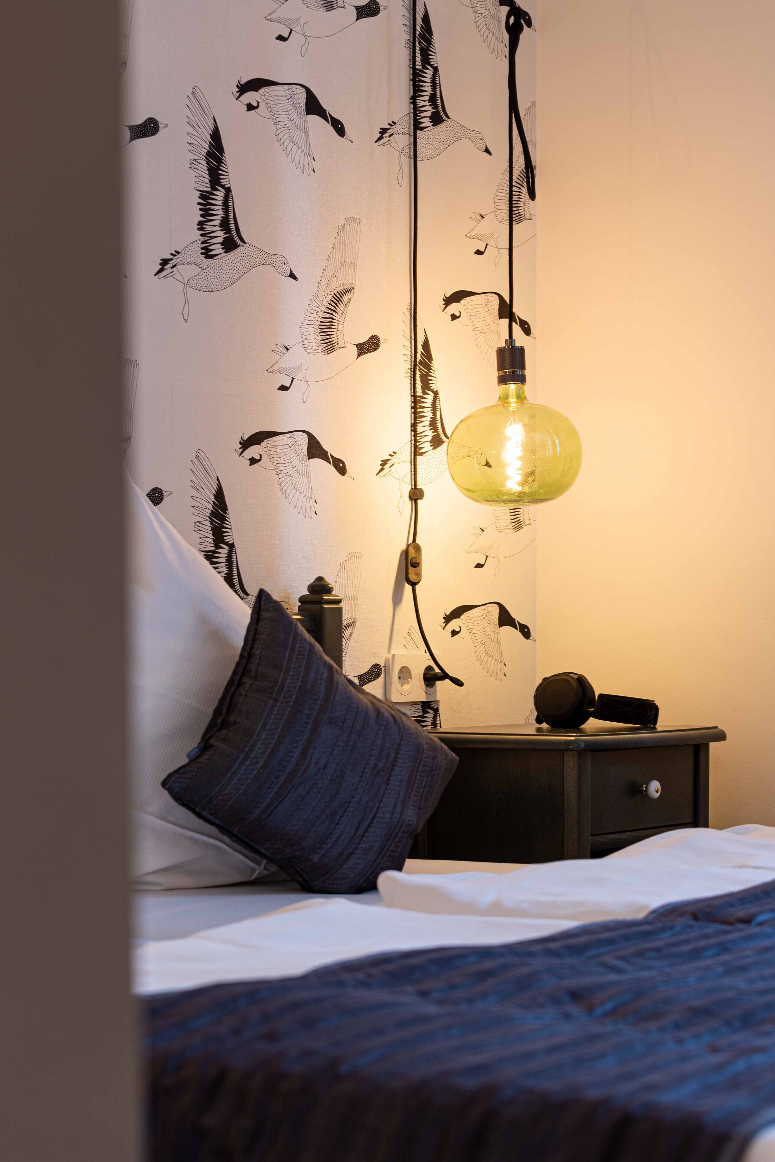 Ein Schlafzimmer mit Bett, Nachttisch, Lampe und einer Wand mit Vogelmuster.