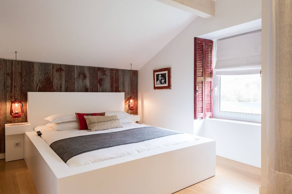 Un dormitorio moderno con una cama grande, un panel de madera en la pared, dos lámparas de noche y una ventana con contraventanas.
