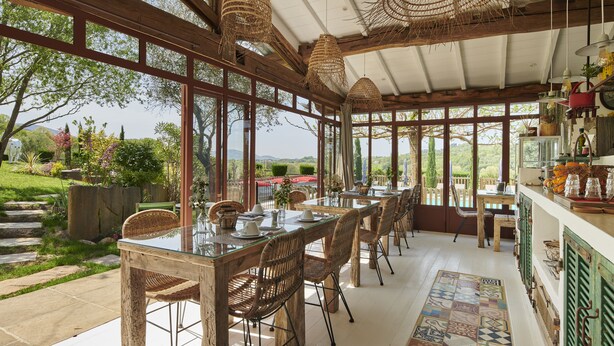 Un área de comedor al aire libre con techo, sillas y mesas de mimbre, ventanas grandes y vistas del paisaje circundante.