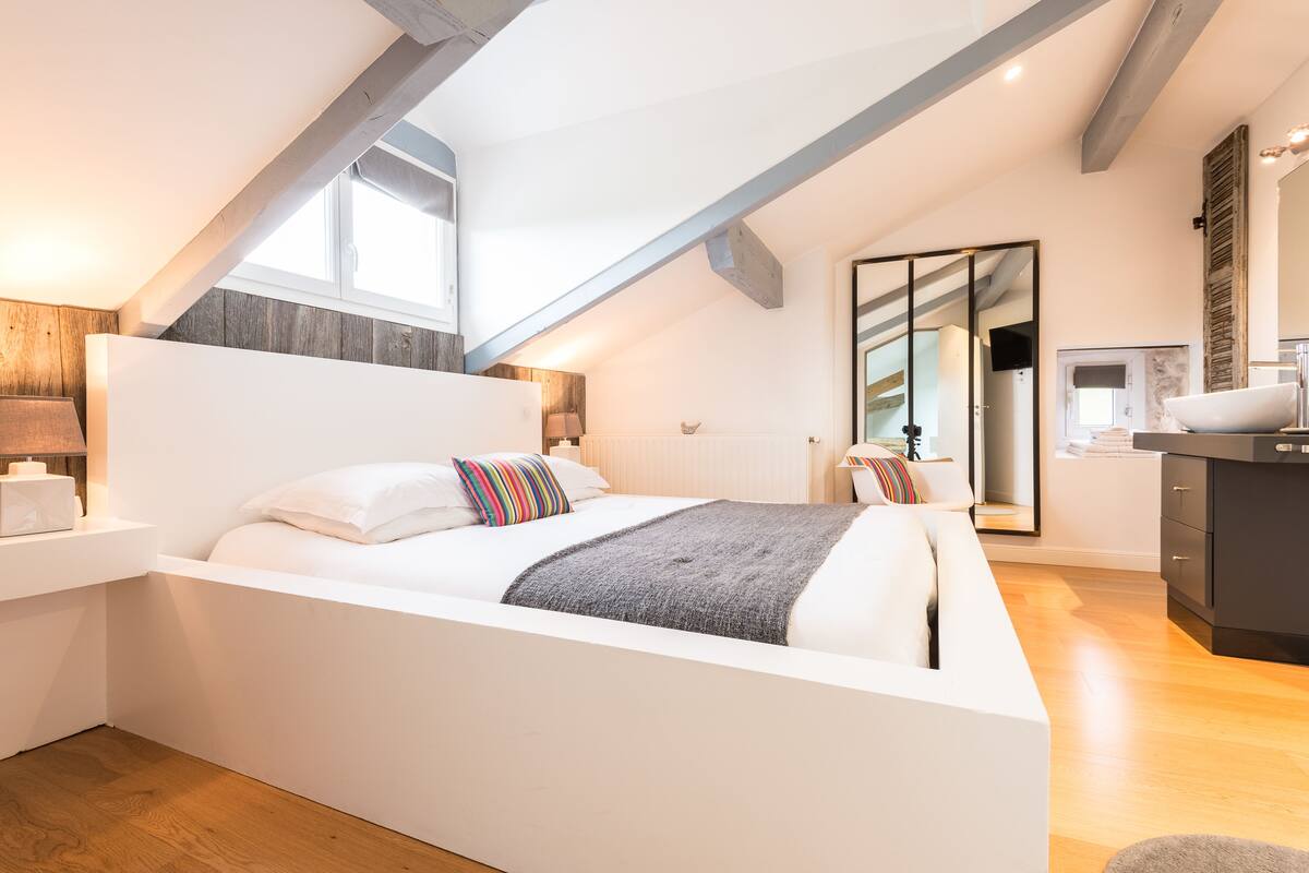 Un dormitorio moderno con una cama grande, vigas de madera, un escritorio pequeño y un baño con espejo y lavamanos.
