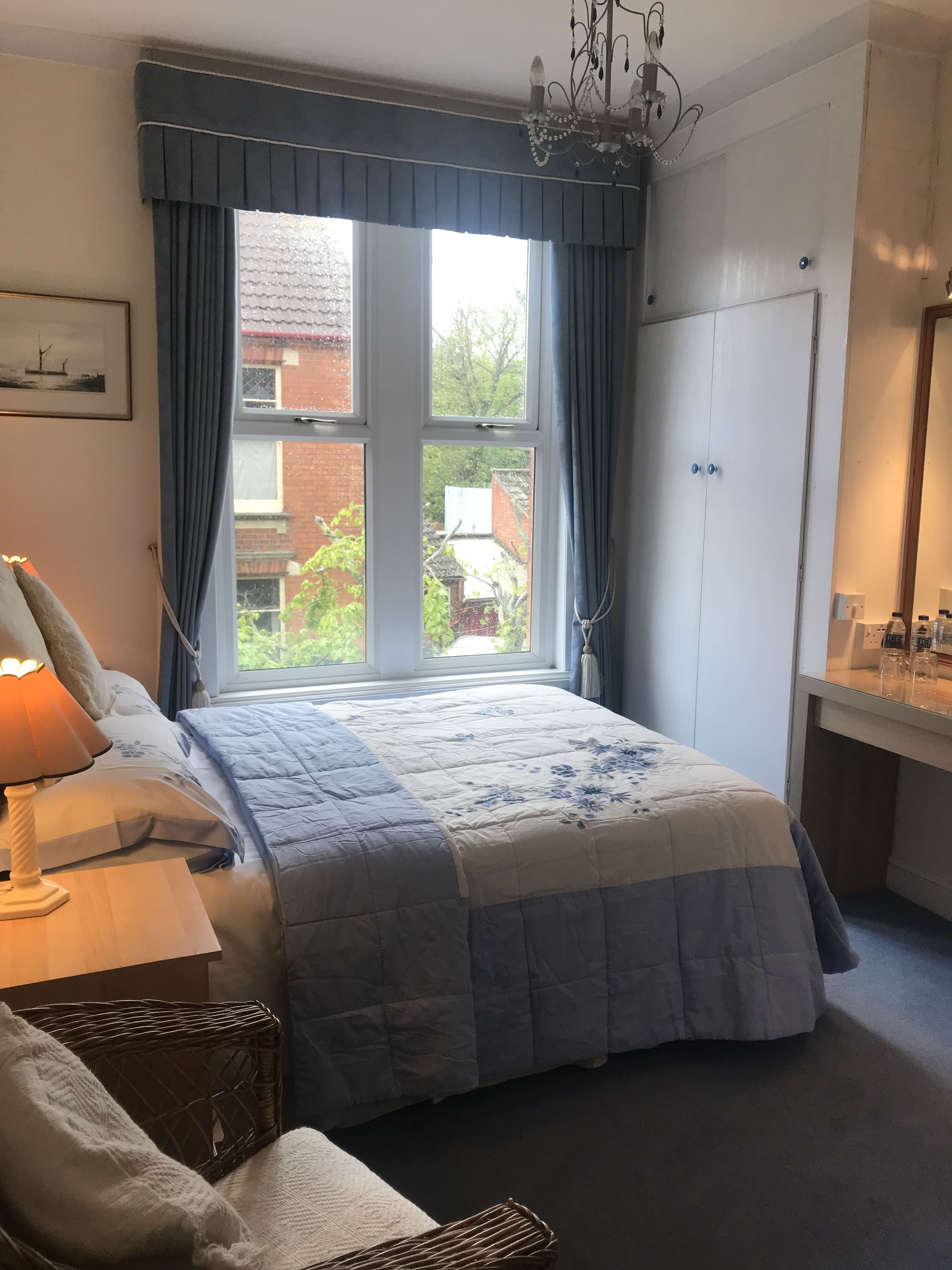 A bedroom with a large bed, a bedside table with a lamp, a wicker chair, and a wardrobe.