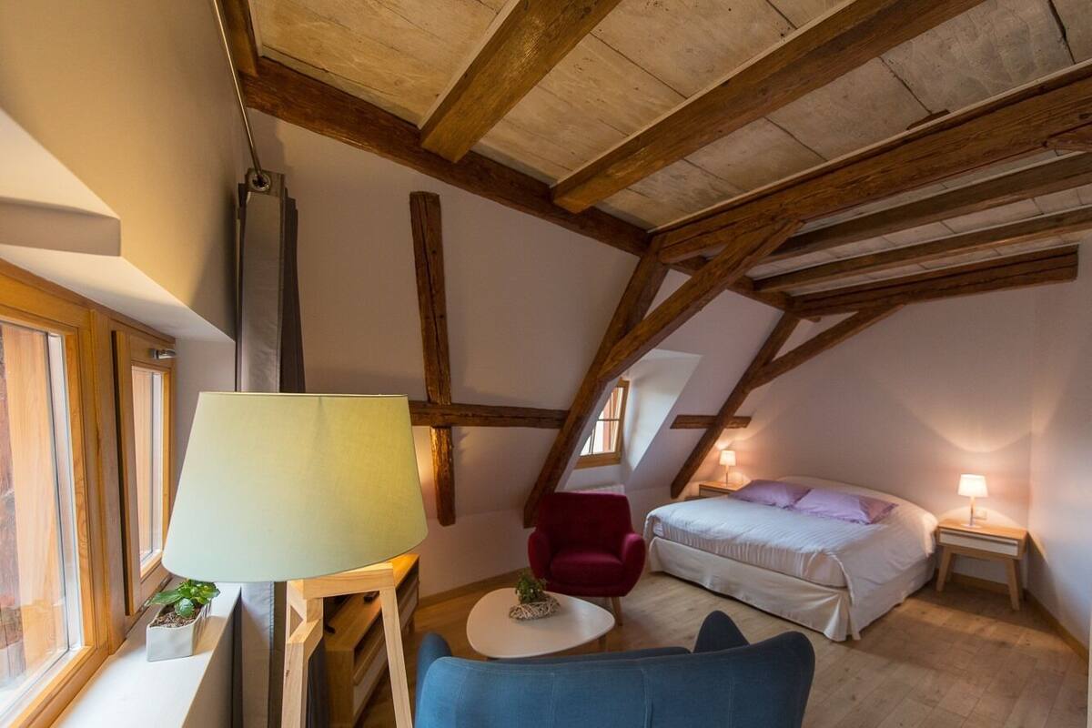 A cozy attic bedroom with a bed, a red chair, a blue sofa, and a small table.