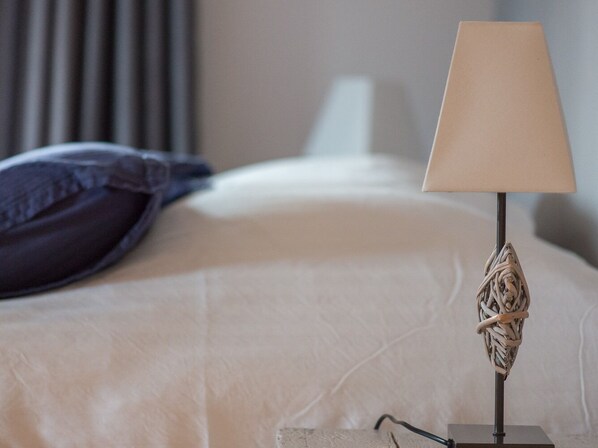 A neatly made bed with a white comforter and a dark blue pillow. A lamp with a woven base is placed on a bedside table.