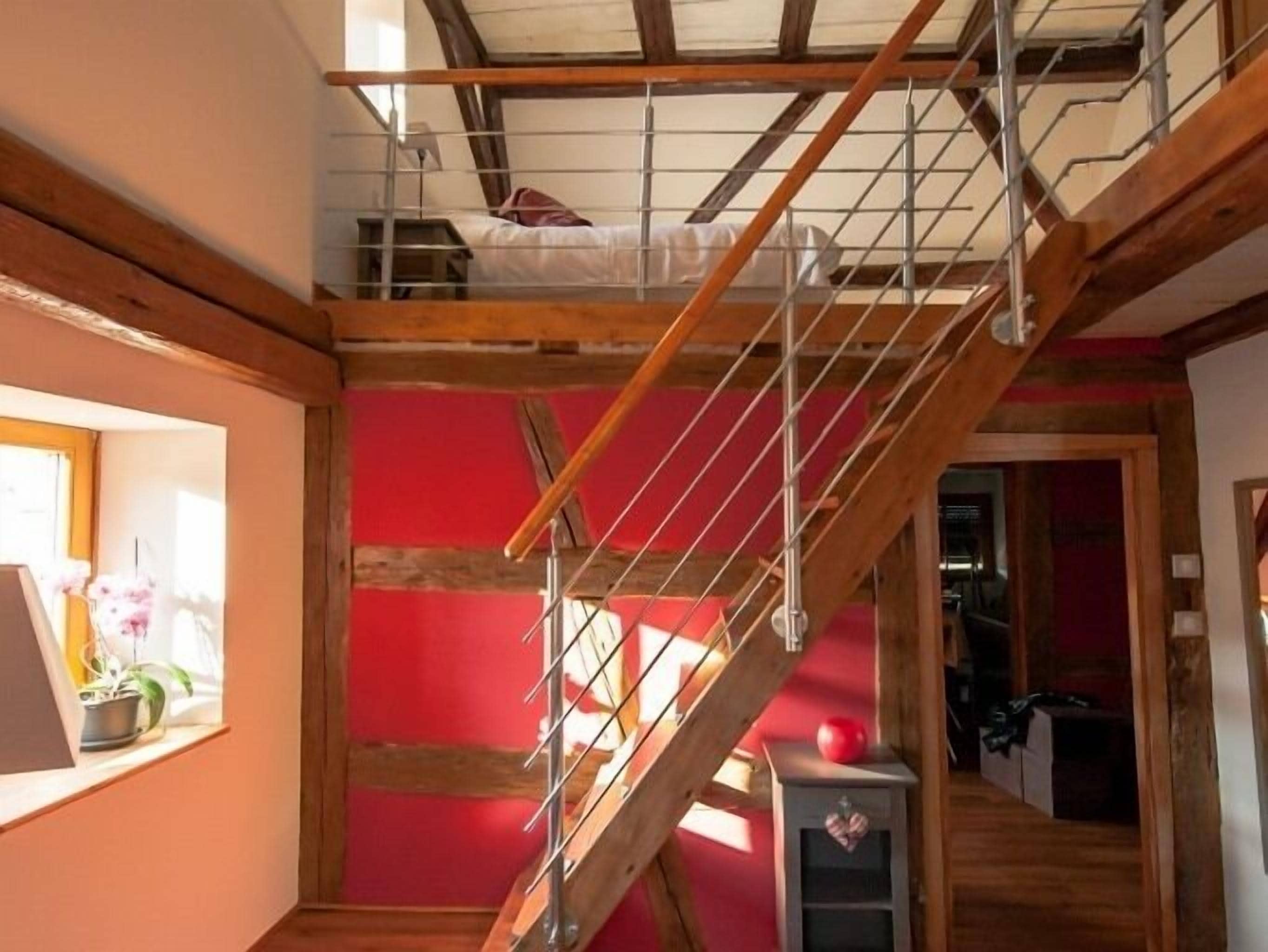 Een kamer met een houten trap, een bed op de bovenverdieping, en een klein tafeltje met een rood voorwerp erop.