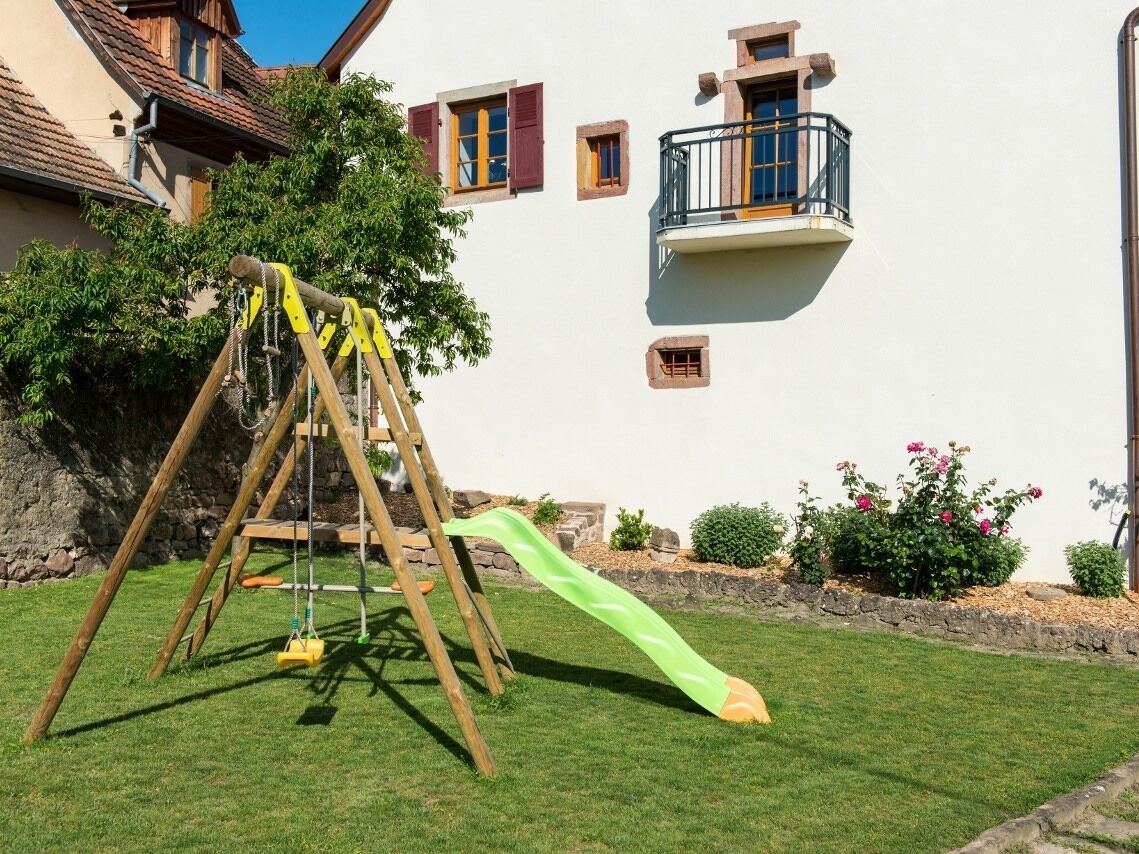 Een houten schommel met een groene glijbaan in een gazon, een stenen muur en een gebouw met een balkon.