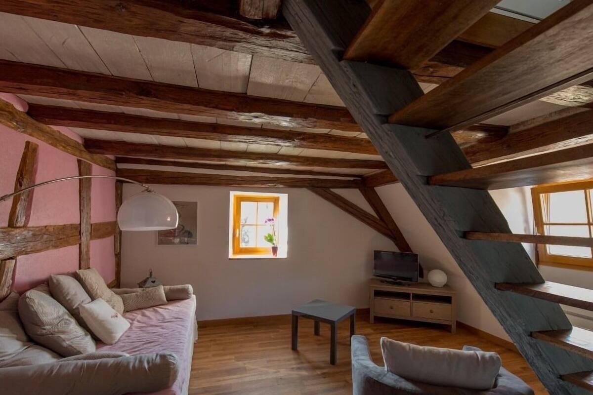 A cozy attic room with exposed wooden beams, a pink sofa, a grey armchair, a small table, and a TV on a wooden stand.