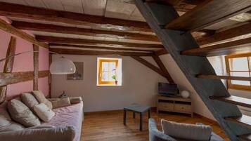 Un acogedor dormitorio en el ático con vigas de madera a la vista, un sofá rosa, un sillón gris, una mesita pequeña y un televisor sobre un soporte de madera.
