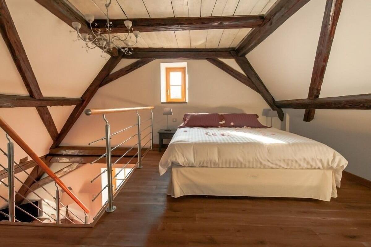 A bedroom with a wooden ceiling, a bed with white and pink bedding, a nightstand, and a window with natural light.