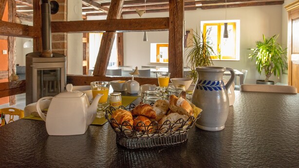 A rustic kitchen with exposed wooden beams, a wood-burning stove, and a breakfast setup including croissants, a teapot, and orange juice.