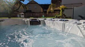 A hot tub with water flowing, surrounded by a stone wall, with houses and a tree in the background.