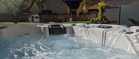 A hot tub with water flowing, surrounded by a stone wall, with houses and a tree in the background.