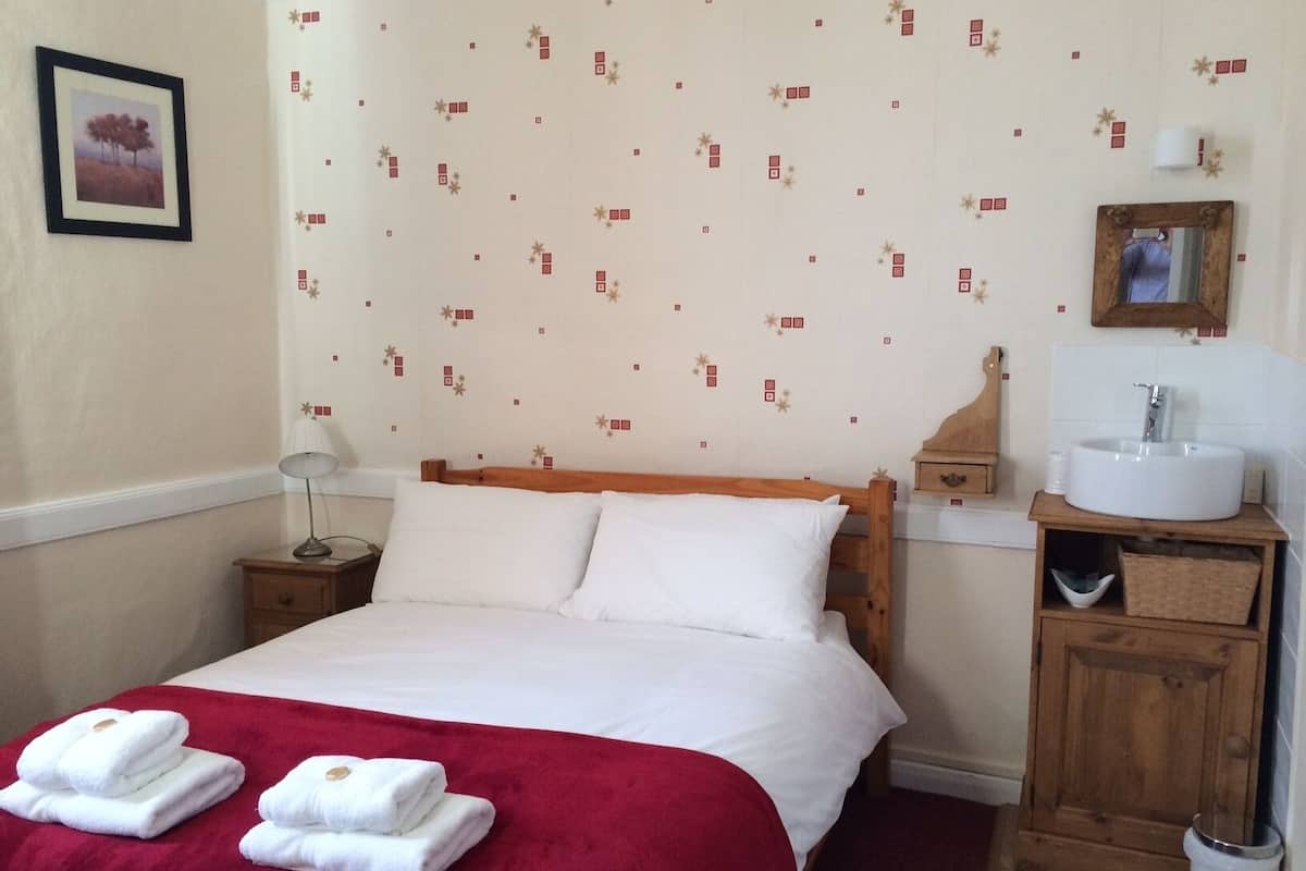 Habitación de hotel con una cama grande, un cabecero de madera, una mesita de noche con lámpara, un mueble de madera con lavamanos y una pared con papel tapiz estampado.
