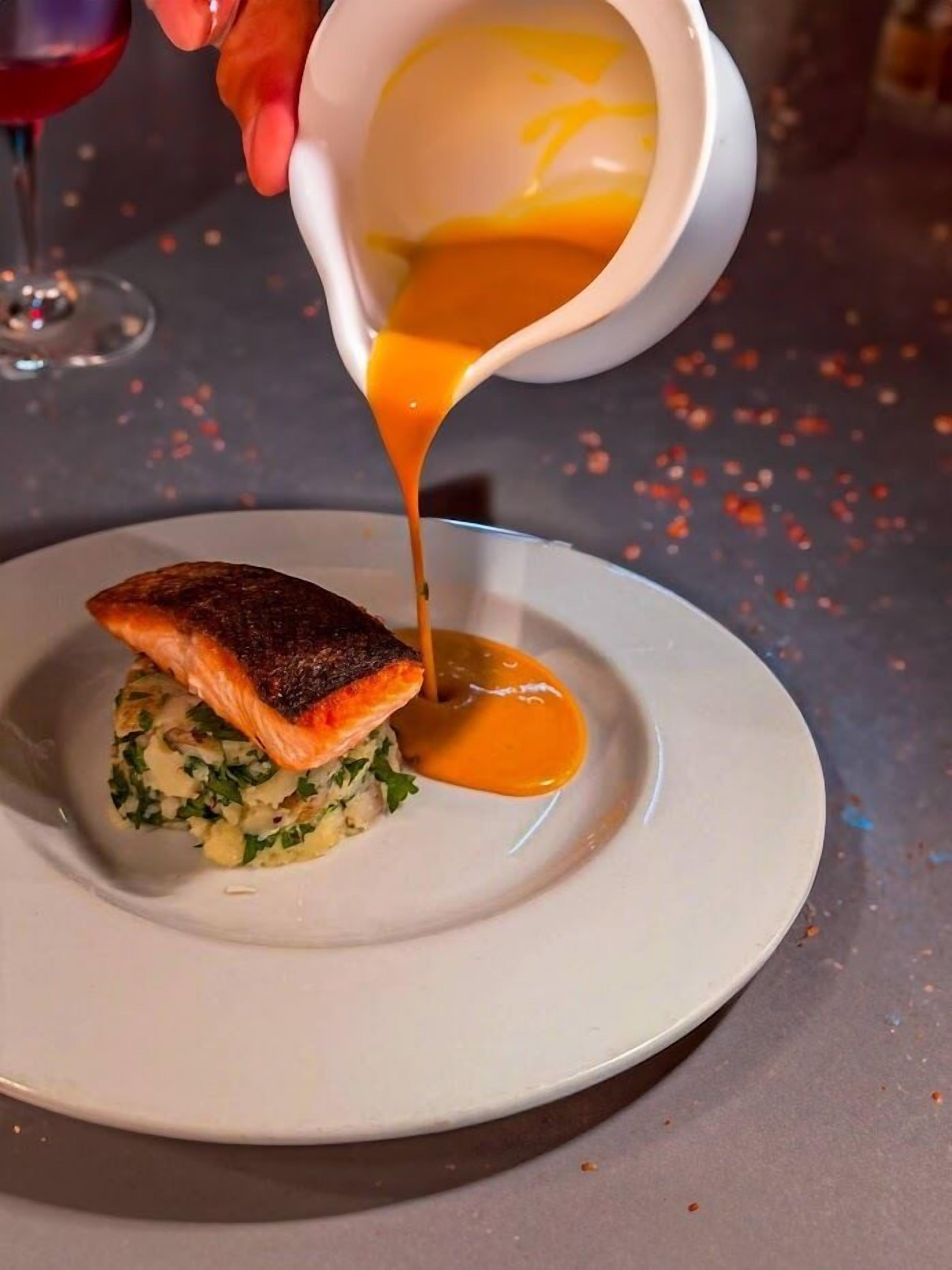 A dish with a piece of salmon on top of a bed of greens, being drizzled with a yellow sauce.