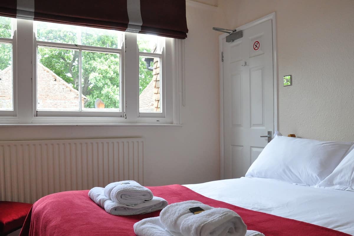 Una habitación de hotel con una cama, un ventanal con vistas a árboles, un radiador y una puerta con un cartel de 'prohibido el acceso'.