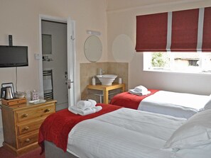 A hotel room with two beds, a TV, a wooden dresser, a bathroom with a sink and mirror, and a window with red curtains. - The Rose & Crown (Etchingham)