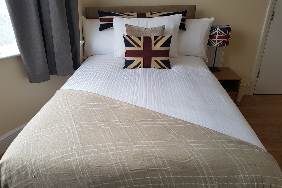 Un lit bien fait, recouvert d’une couette blanche et beige, avec un oreiller orné du motif Union Jack, et une table de chevet sur laquelle se trouve une lampe.
