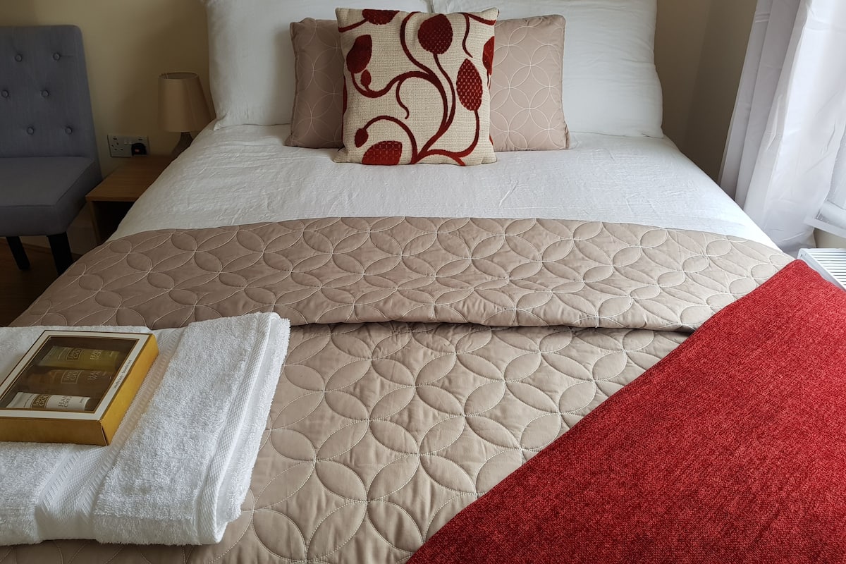 Un lit bien fait, recouvert d’une couette beige, d’un coussin à motifs rouges et blancs, et d’un plaid rouge.