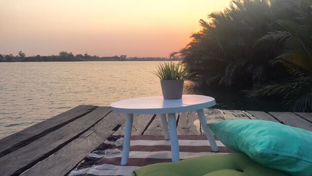 A wooden deck with a white table and a green cushion, overlooking a body of water at sunset. - River Breeze (Chachoengsao)