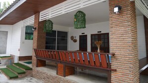 A covered porch with a wooden bench, brick columns, and a small garden area with grass and potted plants. - River Breeze (Chachoengsao)