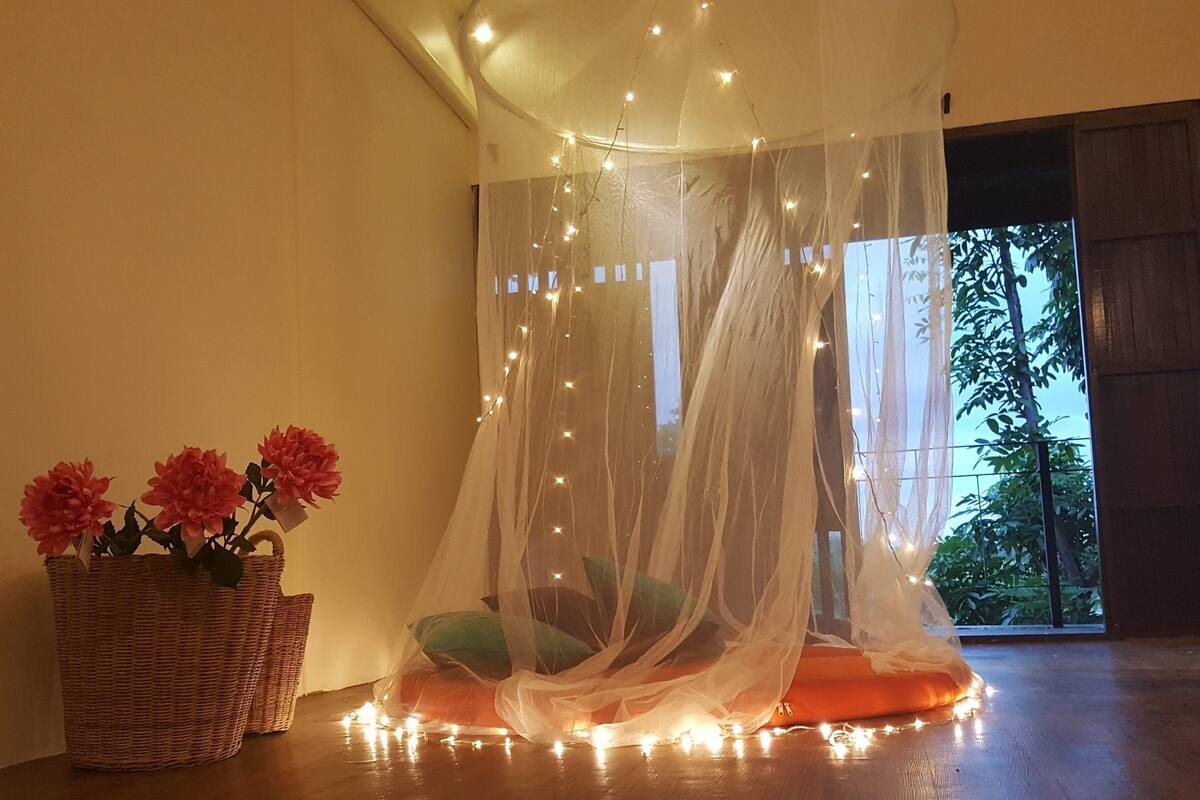 A room with a canopy bed, a wicker basket with flowers, and a view of greenery outside.