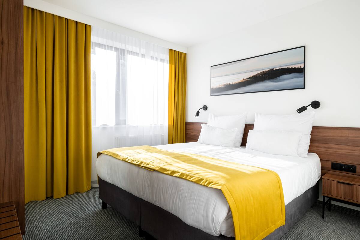 Habitación de hotel con una cama grande, una mesita de noche de madera, un televisor montado en la pared y una ventana con cortinas.